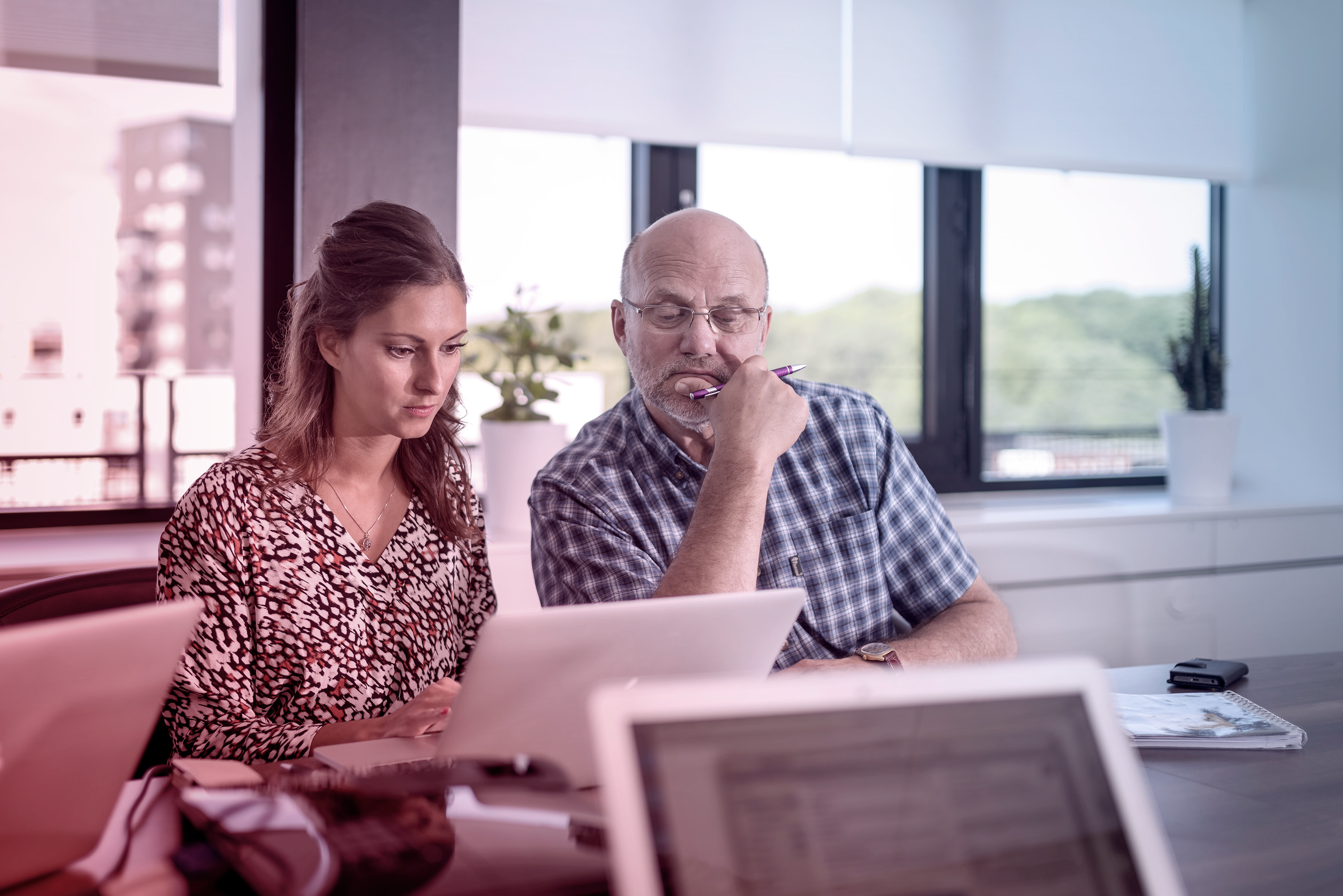 En kvinna och en man sitter sida vid sida vid ett kontorsbord och fokuserar på en dator. Kvinnan pekar lätt på skärmen medan mannen håller en penna mot hakan och funderar. Framför dem står laptops och dokument. Rummet har stora fönster med utsikt över grönska, och växter står på fönsterbrädan. Ljuset är naturligt och stämningen koncentrerad.