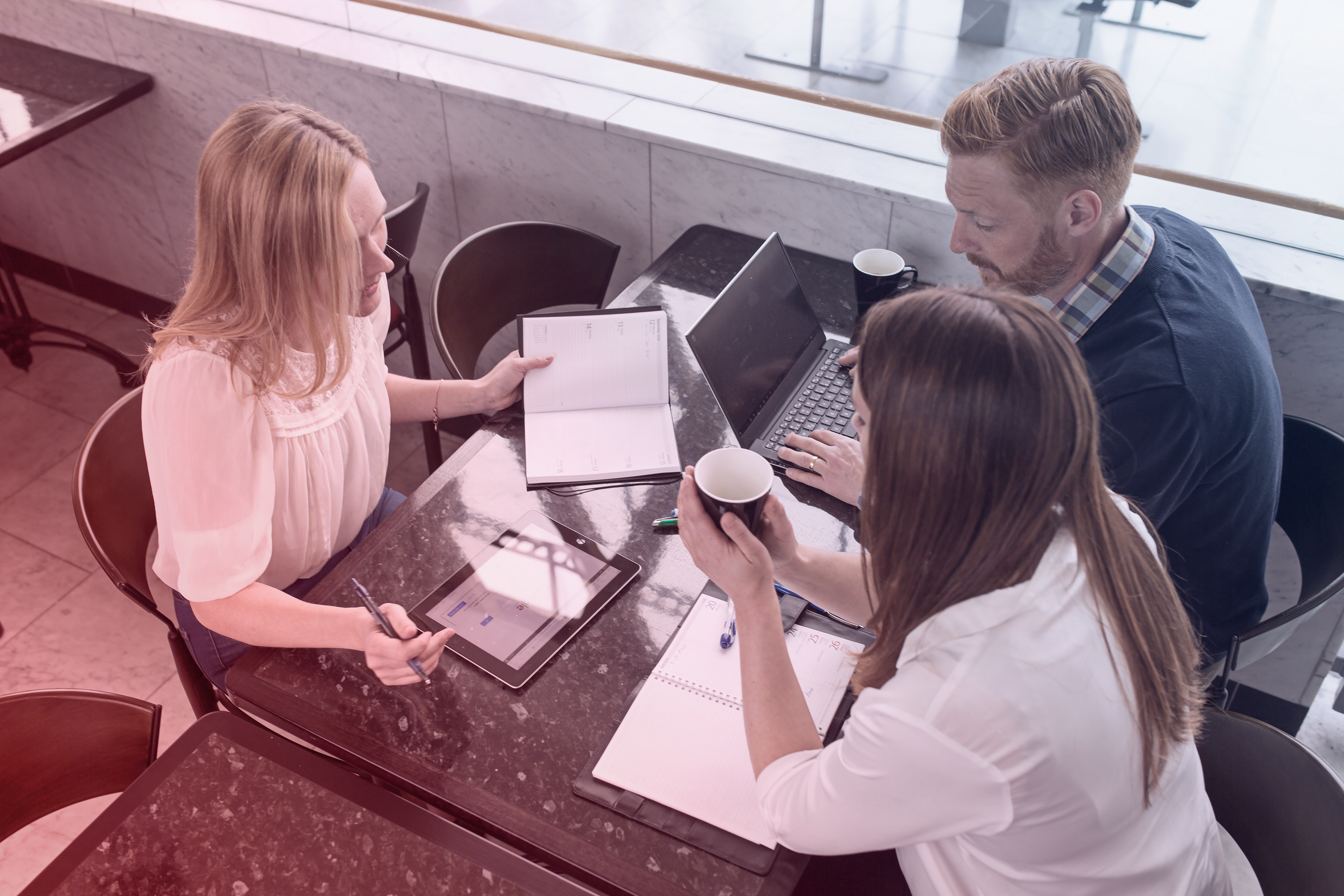 Three professionals sitting at a table in a modern office setting, discussing work with laptops, notebooks, and a tablet.