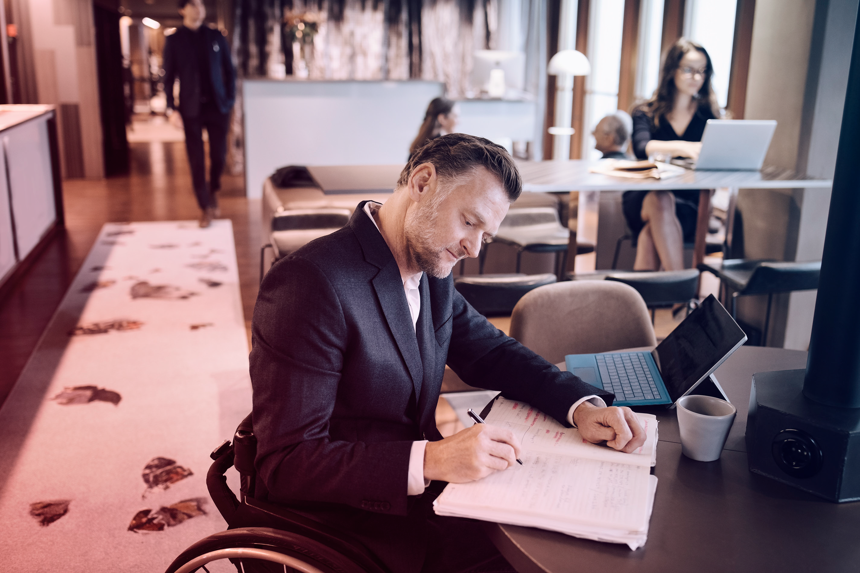 Man in a wheelchair writing in a notebook at a laptop in a modern office space.