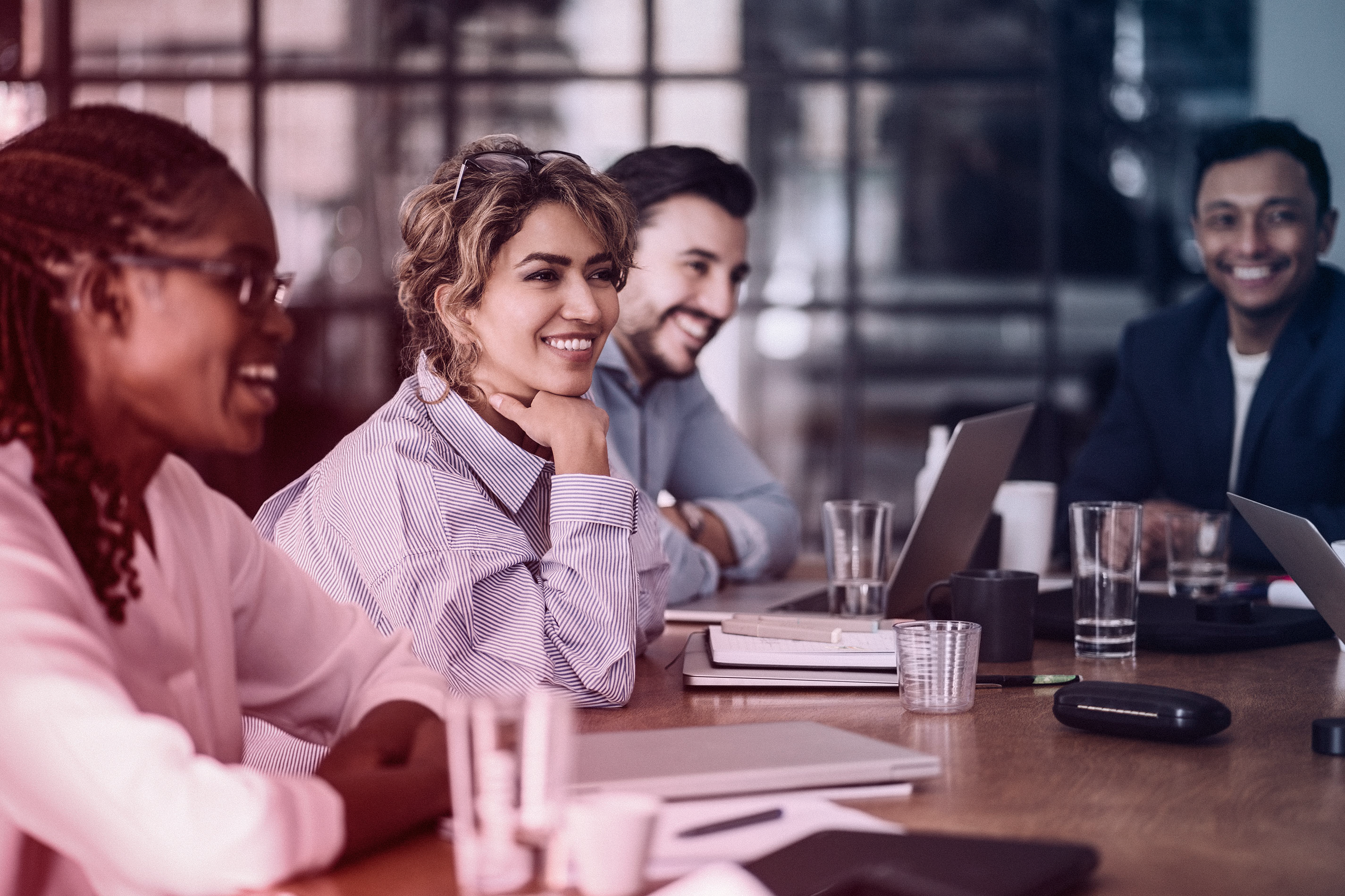 A diverse group of professionals smiling and engaged in a meeting around a conference table with laptops, notebooks, and glasses of water.