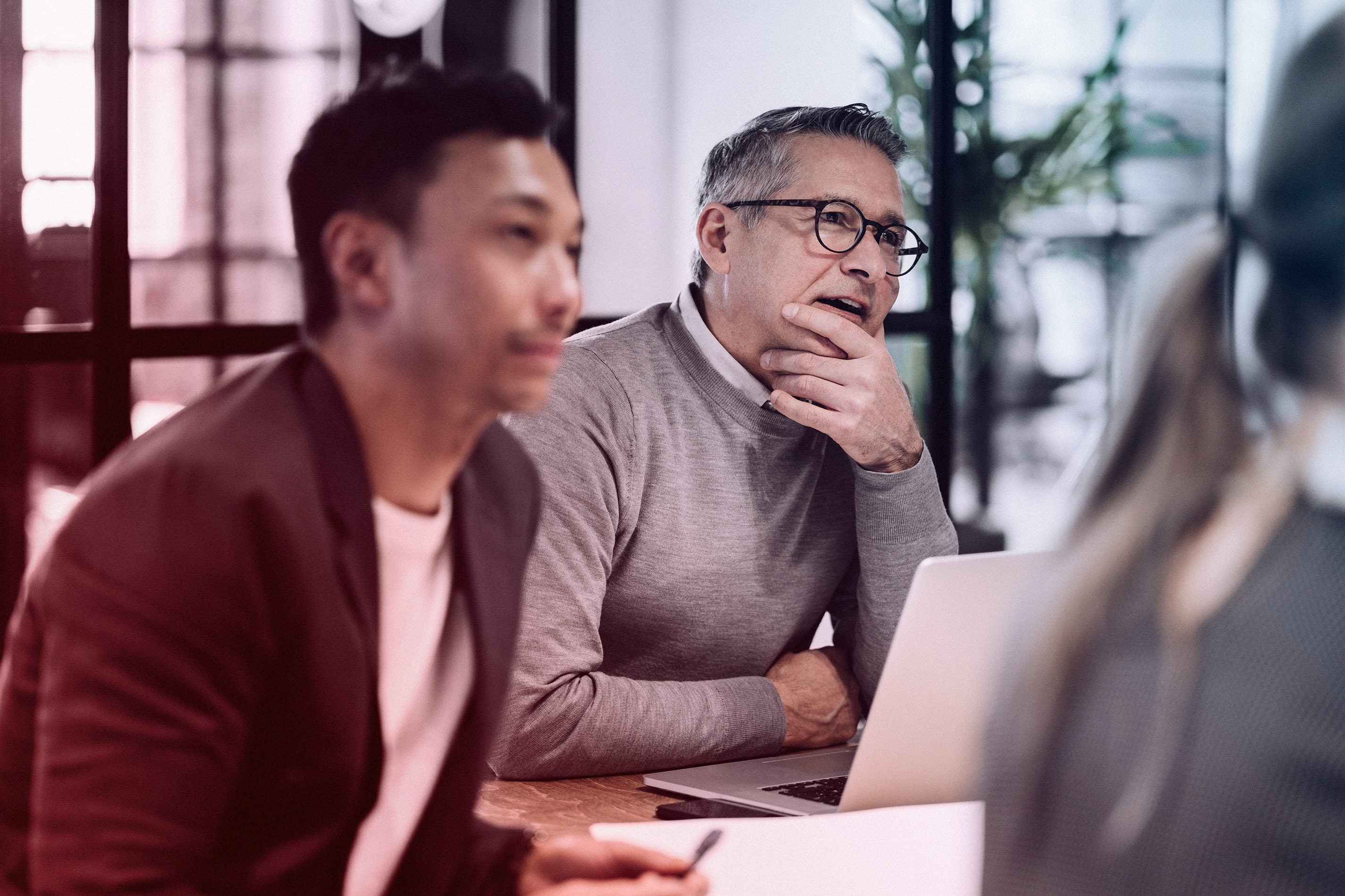 Man in glasses listening thoughtfully during a meeting, seated at a table with a laptop and colleagues.